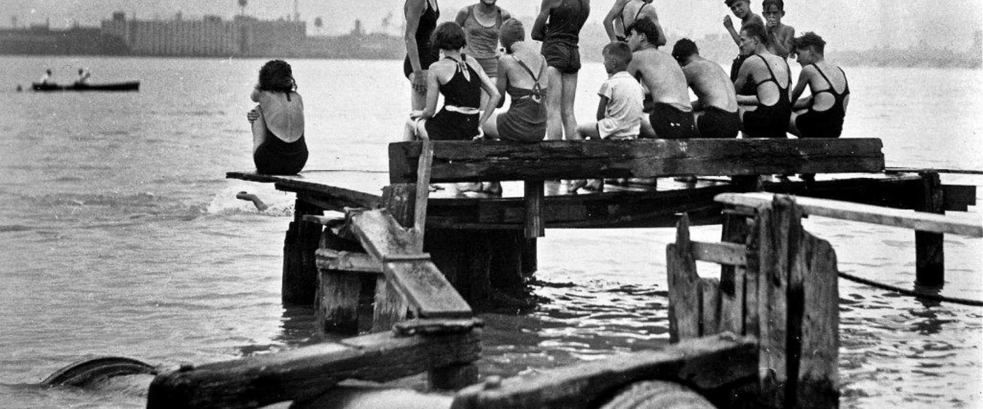 Black and white photo of individuals in their bathing suits siting on makeshift docks, industrial outflow pipes in the water below them. 