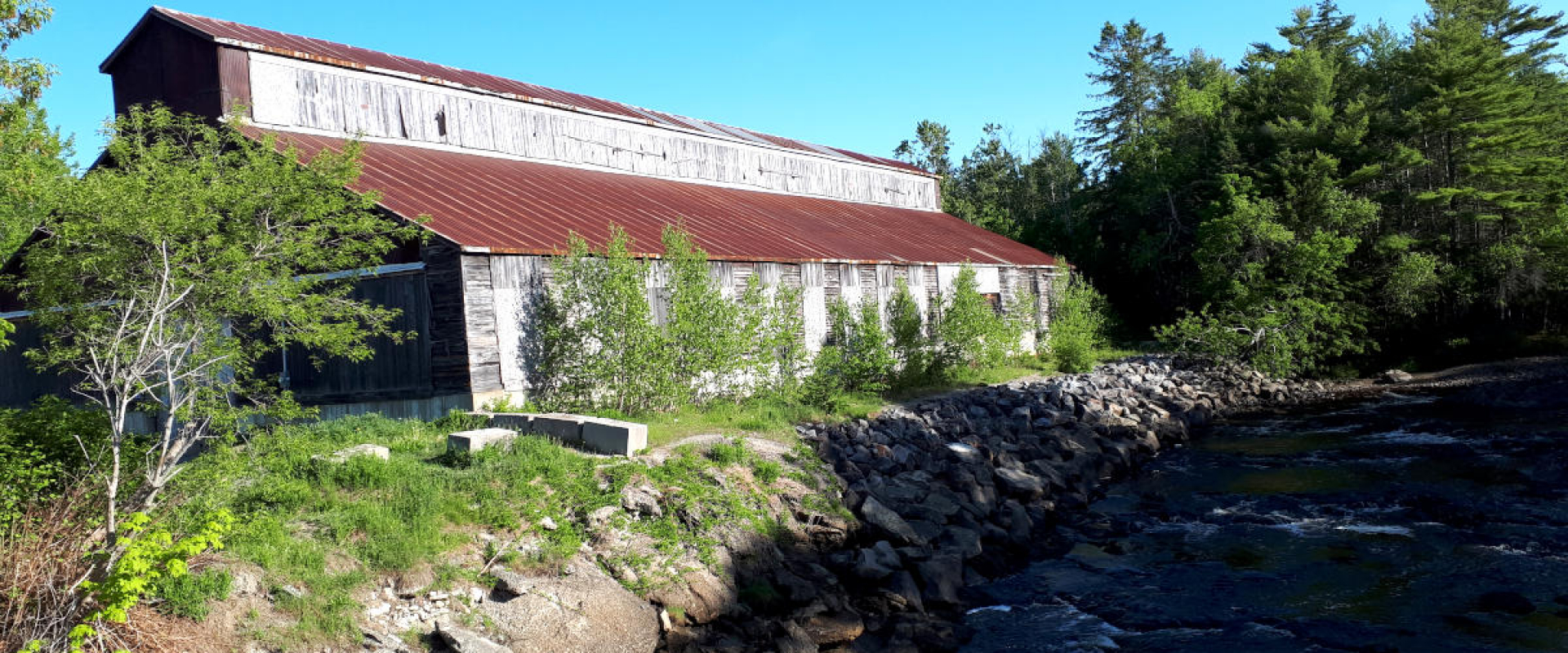 An old, weathered axe factory building that's surrounded by trees on the rocky shore of the St. Croix River. 