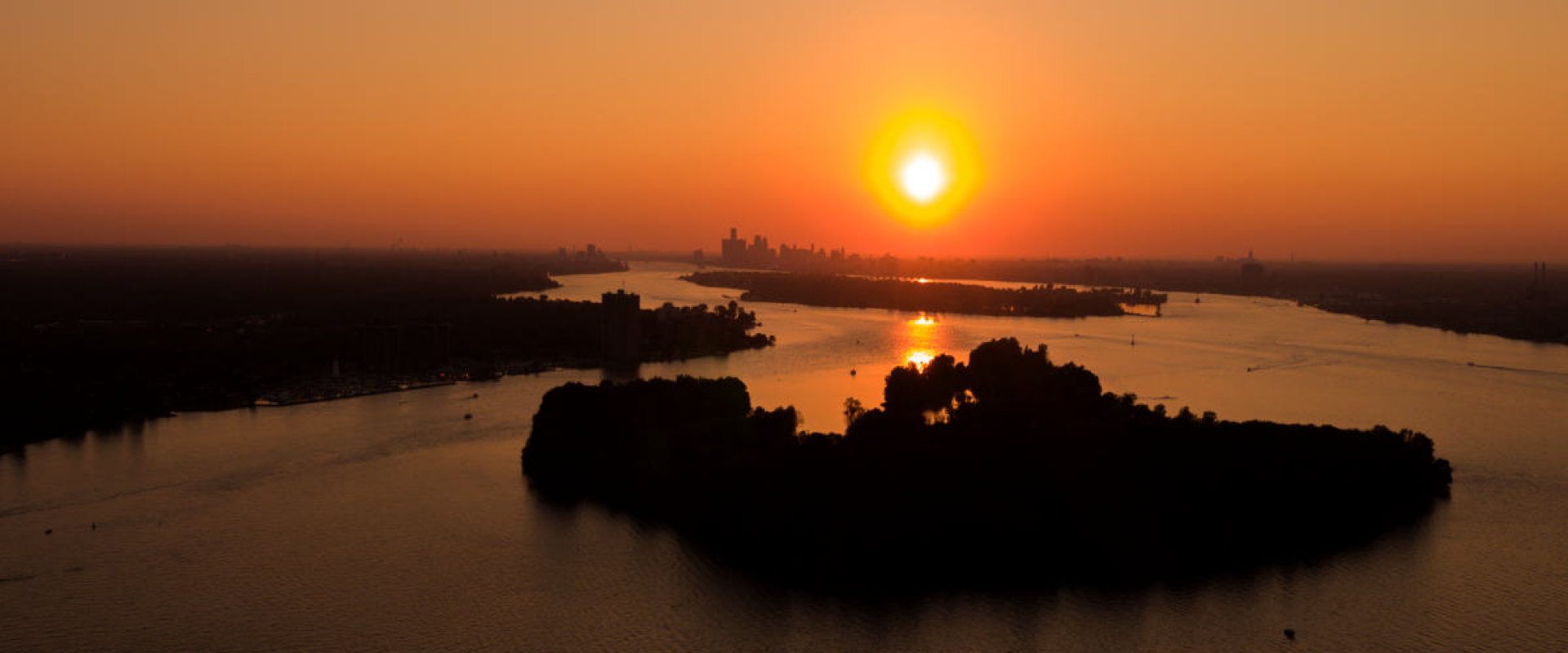The setting sun casts a bright orange glow over Peche Island and the Detroit River