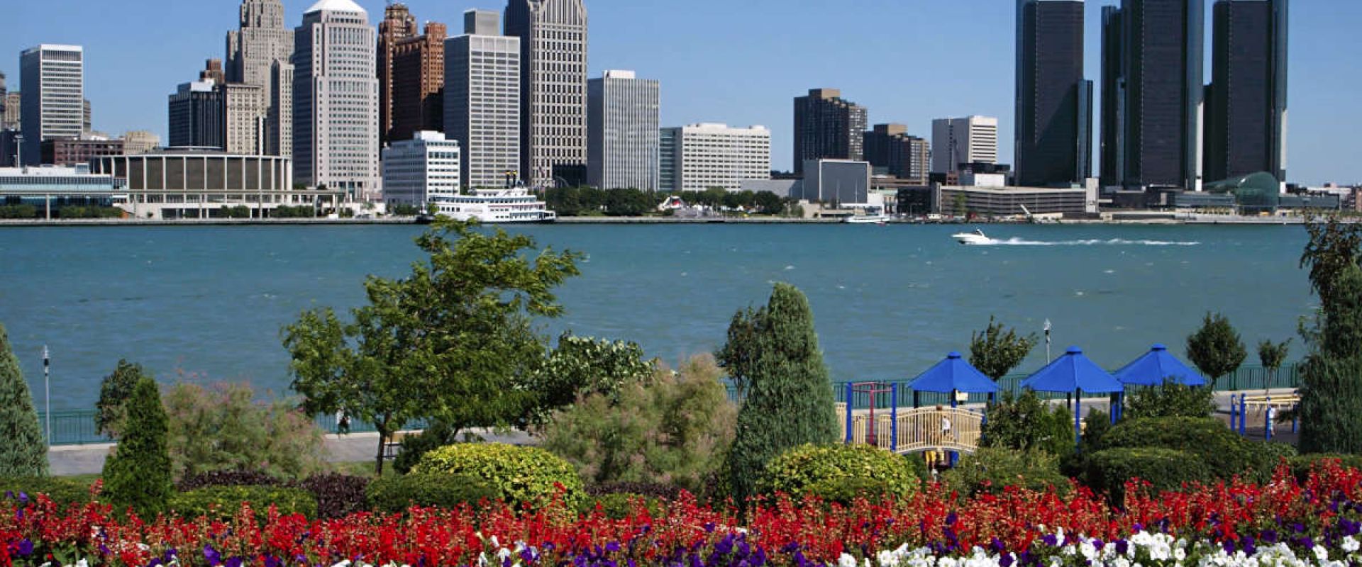 A bed of flowers and a playground nearby the Detroit River on a sunny day. Across the river is an urban landscape that features a collection of skyscrapers.
