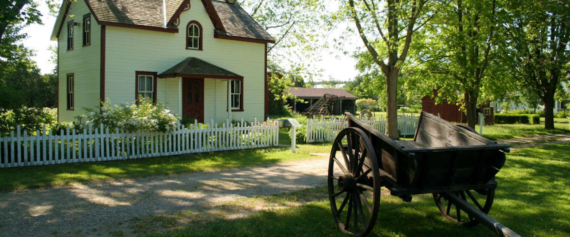 A historic painted wood house with a picket fence and wooden wagon at the Fanshawe Pioneer Village. 