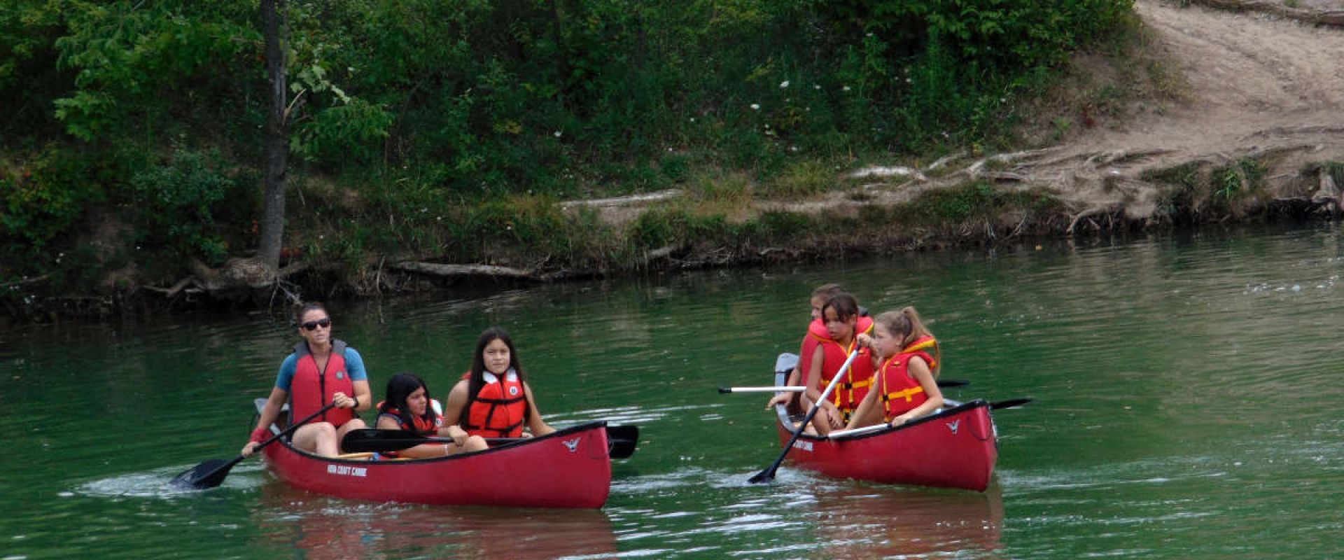 Children accompanied by an adult paddling two red canoes on the Sharon Creek Reservoir. 