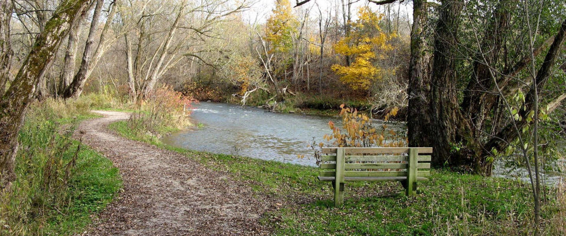 The Woodstock Trail in Burgess Park is a footpath that follows alongside the Thames River. There is a bench pictured for visitors to sit and admire the river.
