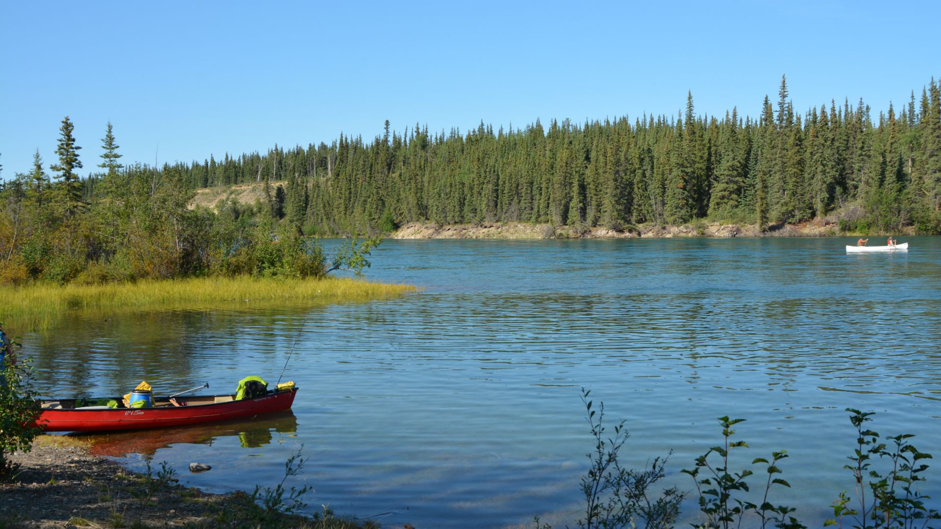 Un canot repose sur la rive de la rivière Thirty-Mile, par une journée ensoleillée, pendant qu'un autre canot avec deux personnes à bord passe au loin.