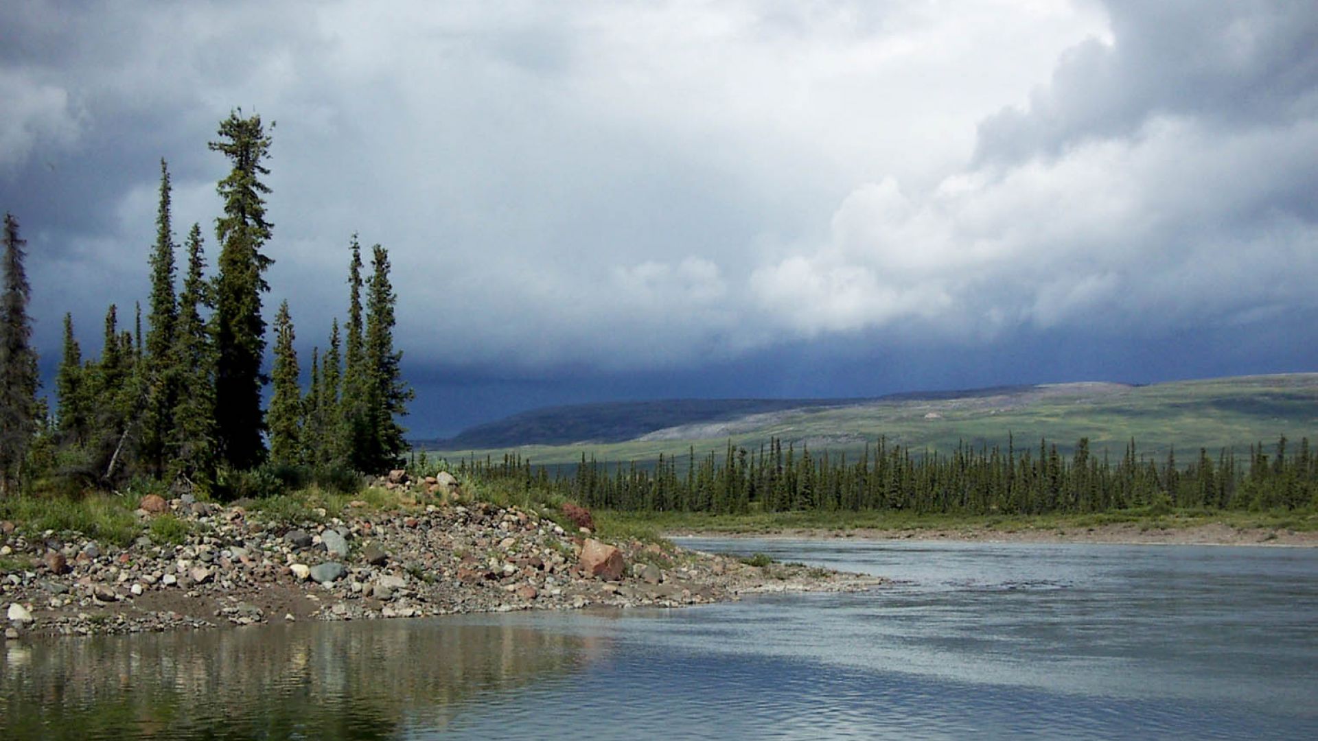 The still water of the Coppermine River reflects the trees and rocks of the shoreline on a cloudy day.