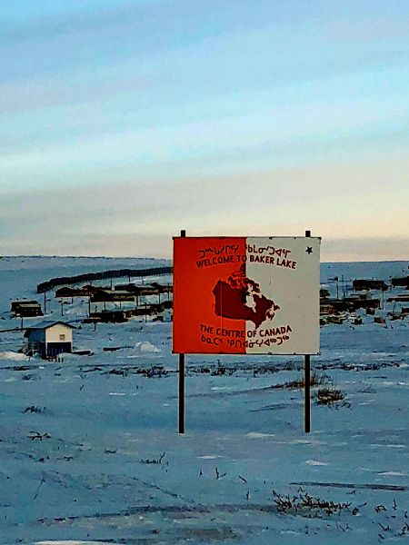 A sign reads out "Welcome to Baker Lake, The Centre of Canada" in both English and Inuktitut. Behind the sign, a small town is visible on the snowy hills.