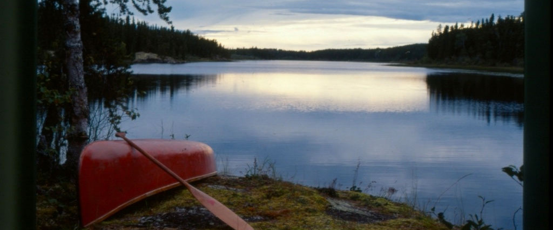 A red canoe and paddle lie near the shoreline of a campsite at dusk.