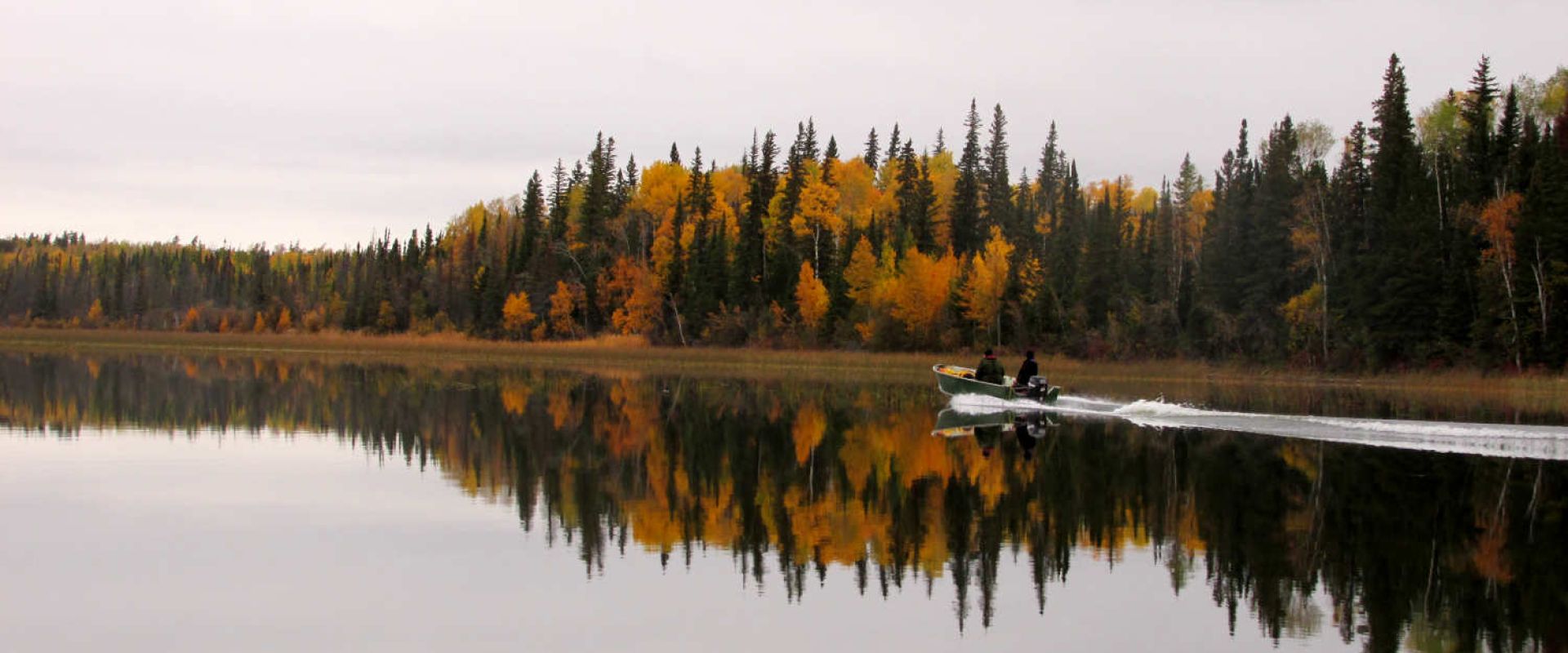 Two people travel by motorboat across a glassy lake that is connected to the Hayes River in the fall.