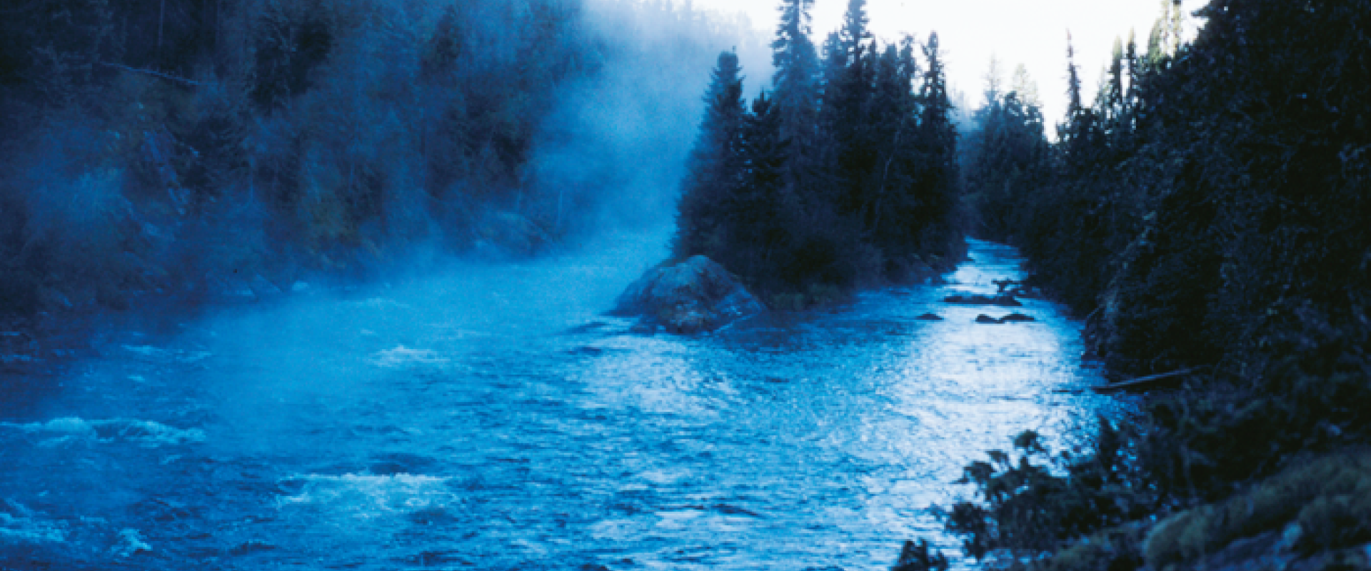 A treed shoreline can be seen along the Hell's Gates Gorge. Mist in the photo makes the lighting appear blue. Rapids can be seen in the water.